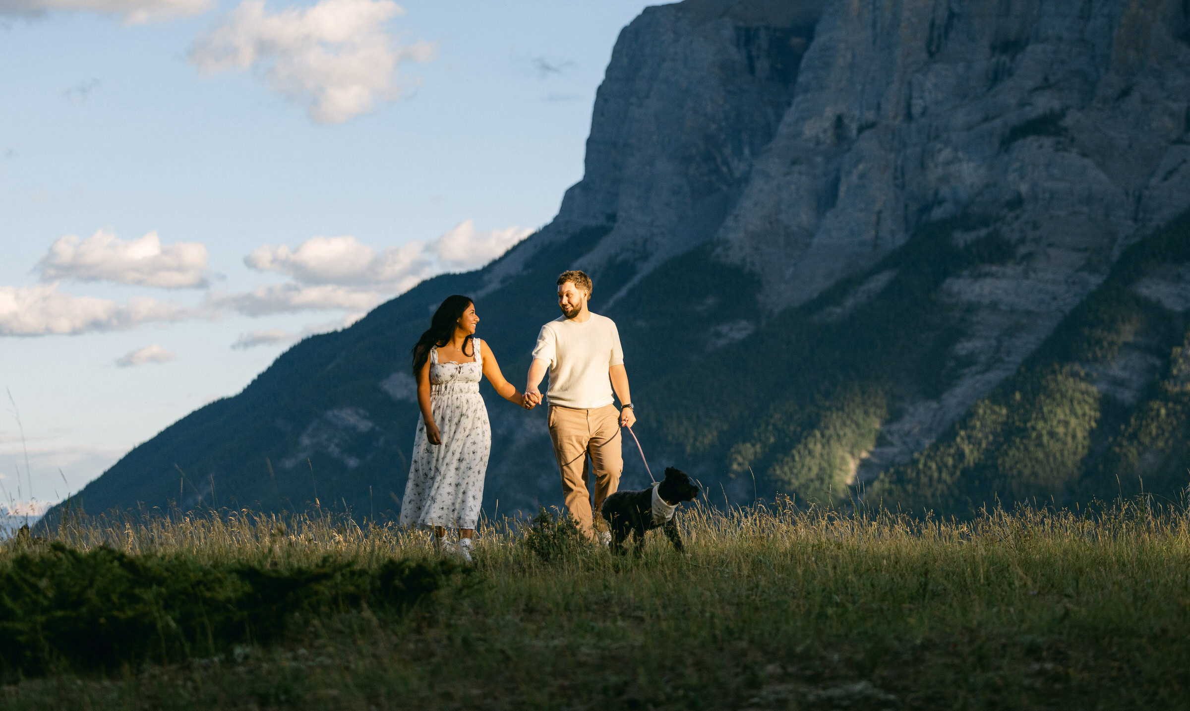 A couple holding hands while walking a dog in a grassy landscape with mountains in the background.