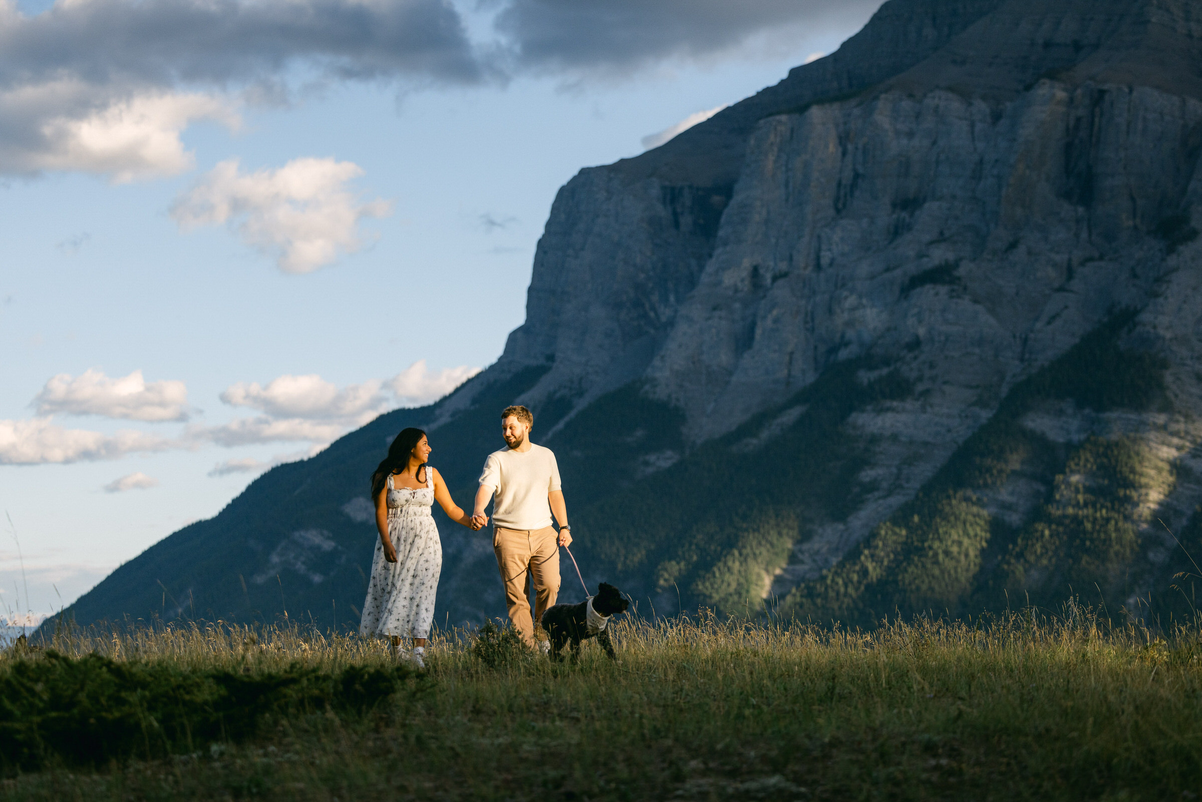 A man and woman holding hands and walking with their dog in a field with a backdrop of towering mountains and a clear sky.