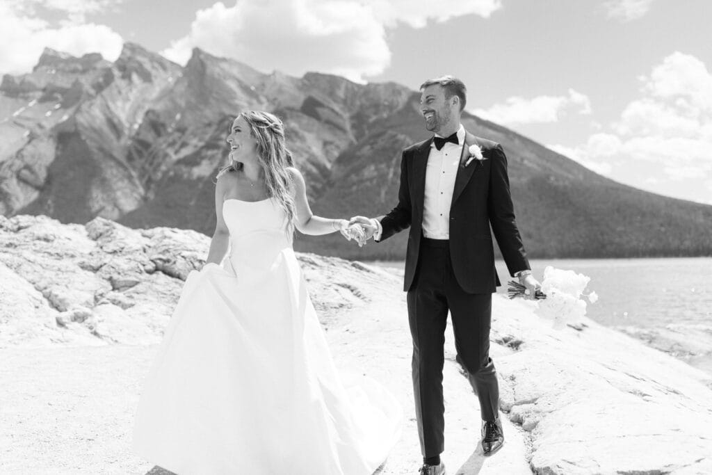 A joyful couple dressed in wedding attire walking hand-in-hand along a rocky shoreline with mountains in the background.