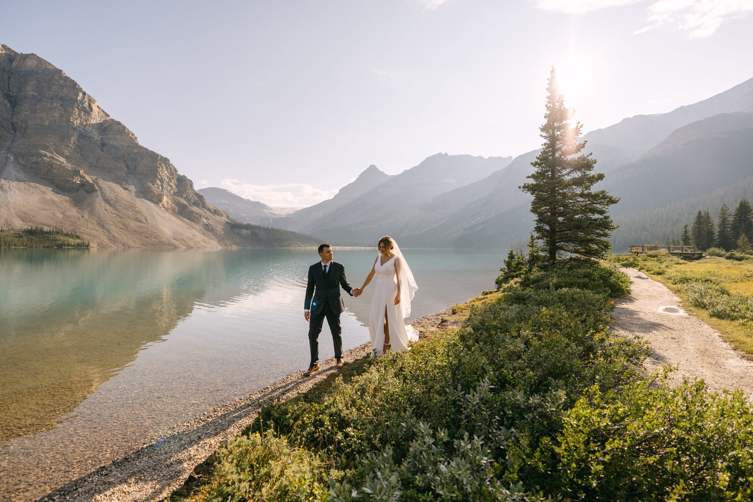 A newlywed couple walks hand-in-hand along a scenic lakeshore, surrounded by mountains and greenery, capturing a beautiful moment of love and serenity.