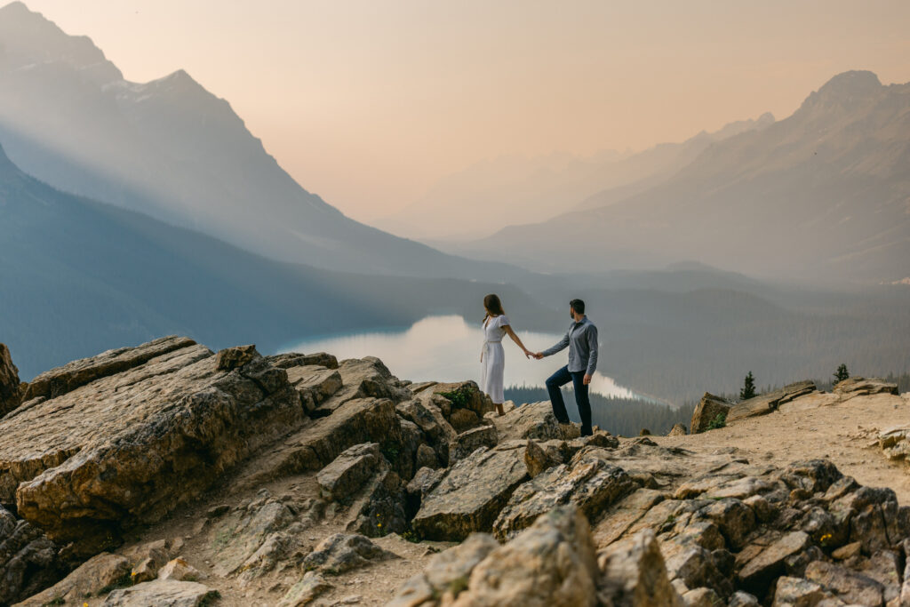 A couple holding hands on a rocky overlook with a hazy mountain sunset in the background.