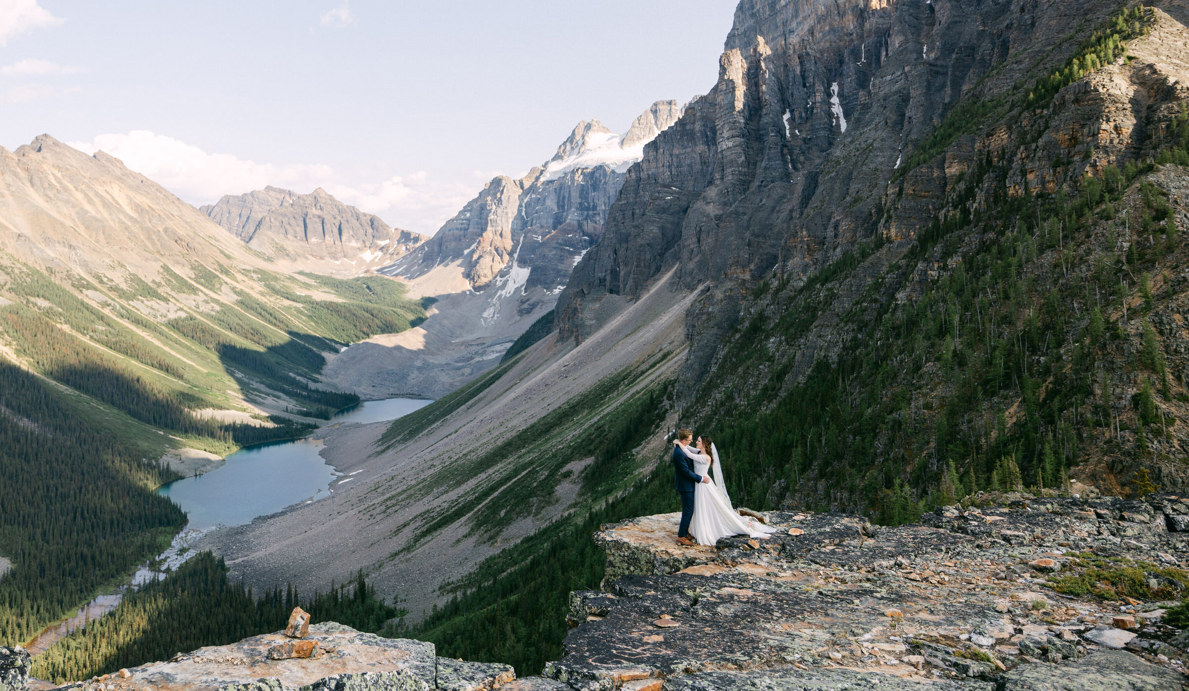 A couple sharing a romantic moment on a rocky ledge overlooking a stunning valley and lake, surrounded by majestic mountains.