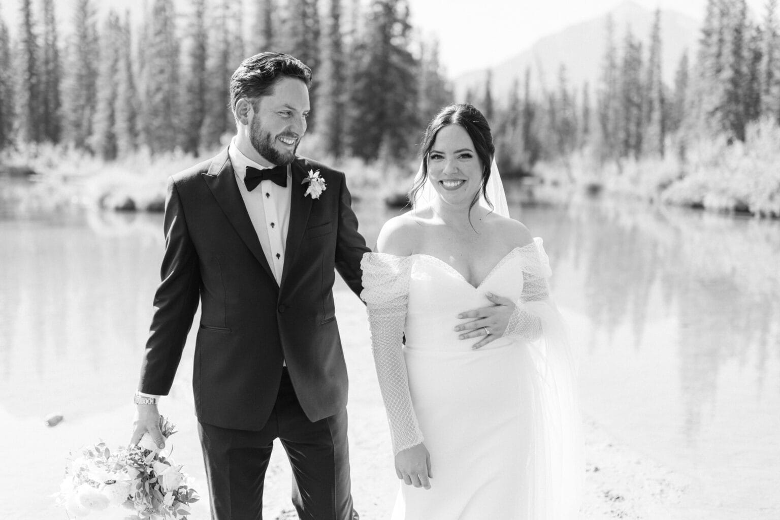 A black and white image of a happy couple on their wedding day, holding hands and smiling, standing by a lake with trees in the background.