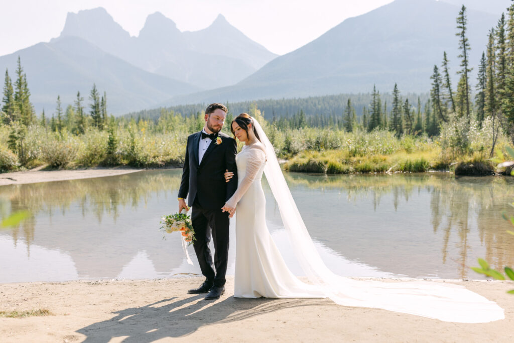 A bride in a long white gown with a trailing veil and a groom in a black suit holding hands by a serene mountain lake.