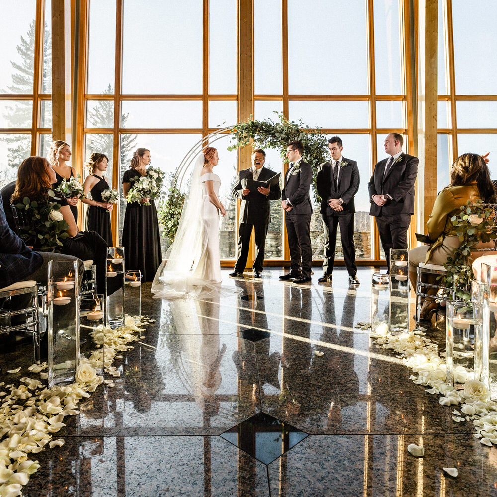 A couple exchanging vows at an indoor wedding ceremony with guests and bridal party looking on, featuring a glass wall background and floor covered in flower petals.