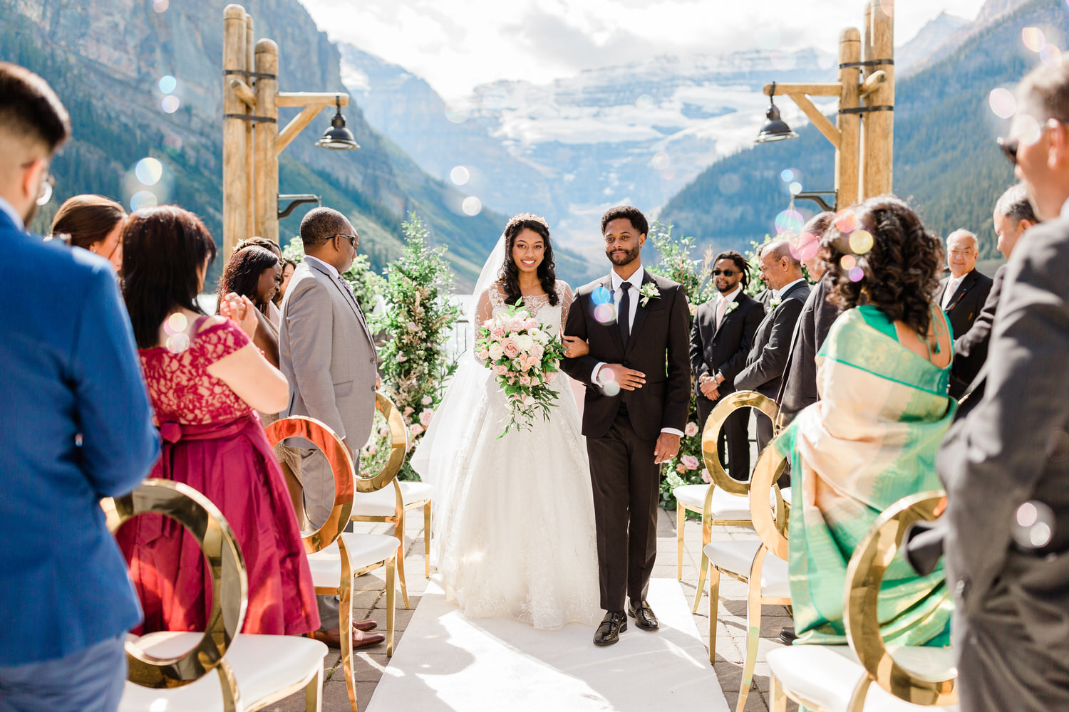 A bride and groom walking down the aisle with guests on either side, set against a backdrop of majestic mountains and clear skies.