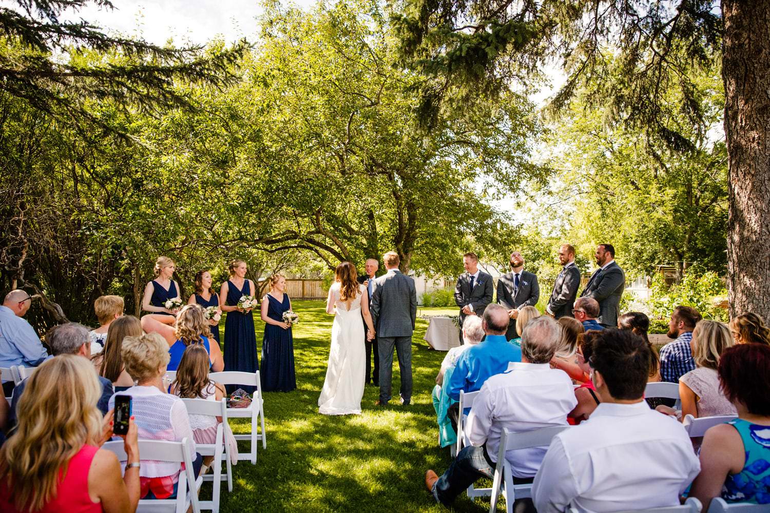 A bride and groom stand before an officiant at an outdoor wedding ceremony, with guests, bridesmaids, and groomsmen around them in a garden setting.