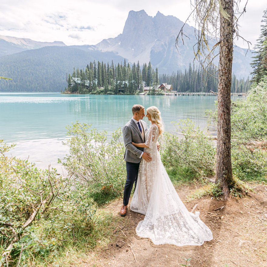 A bride and groom hold each other beside a picturesque mountain lake with a forest and cabins in the background.