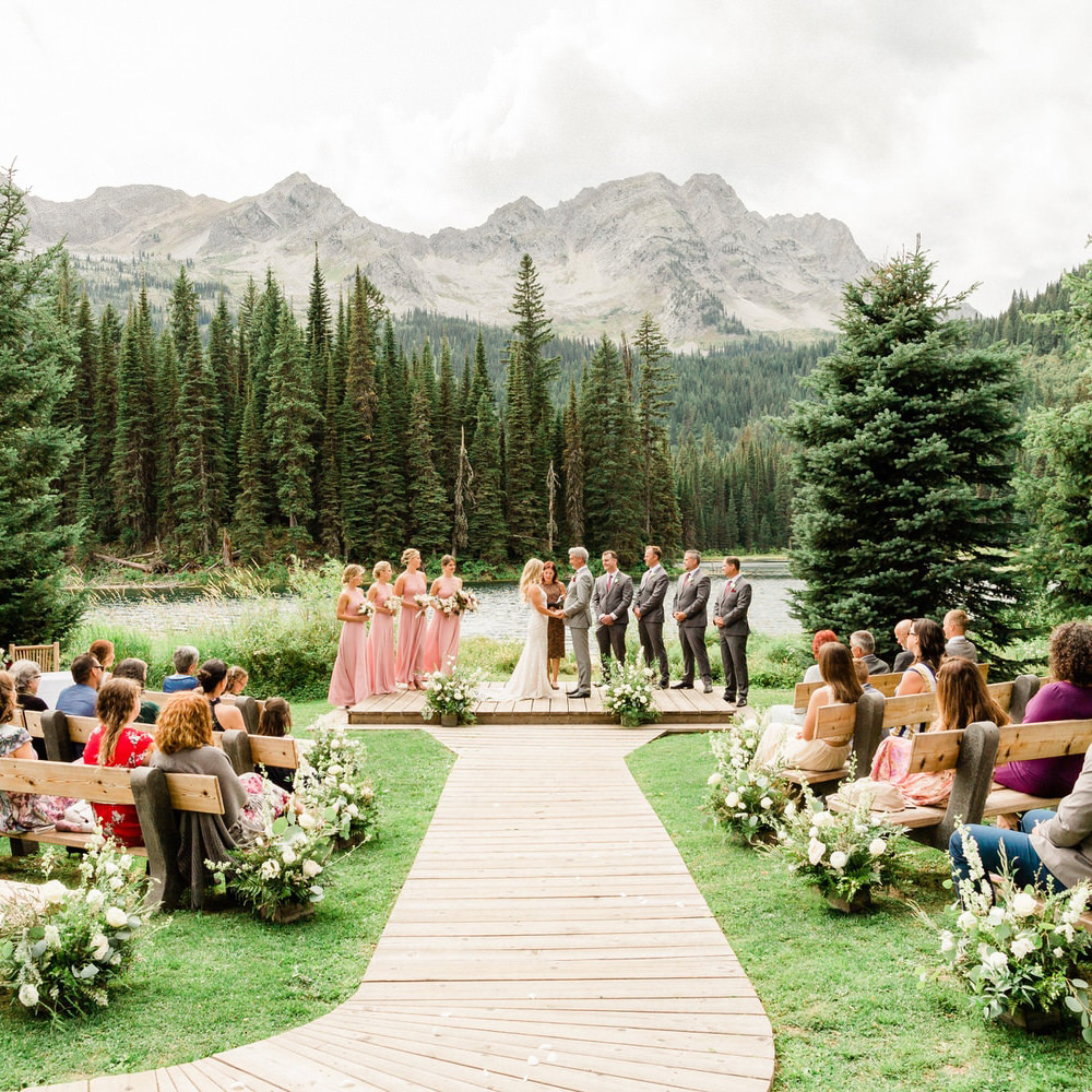 A wedding ceremony taking place outdoors with guests seated on wooden benches, bridal party in pink dresses, groomsmen in suits, and a mountainous backdrop, surrounded by a pine forest.