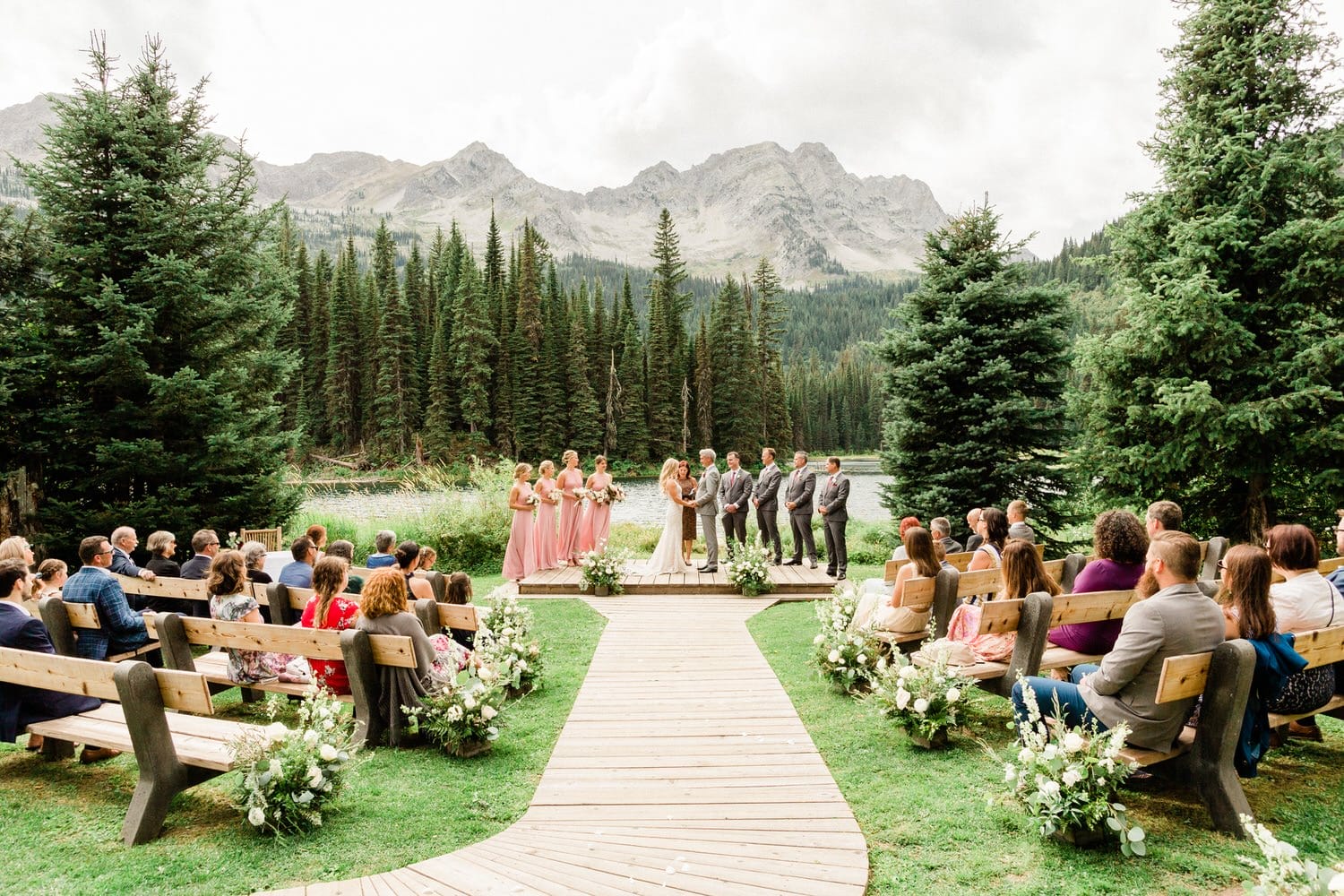 Guests seated on wooden benches at an outdoor wedding ceremony with a bridal party standing in front of a mountainous forest backdrop.