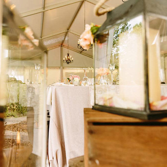 A beautifully decorated wedding reception table under a tent with hanging flowers and chandeliers, viewed through a reflective glass surface.