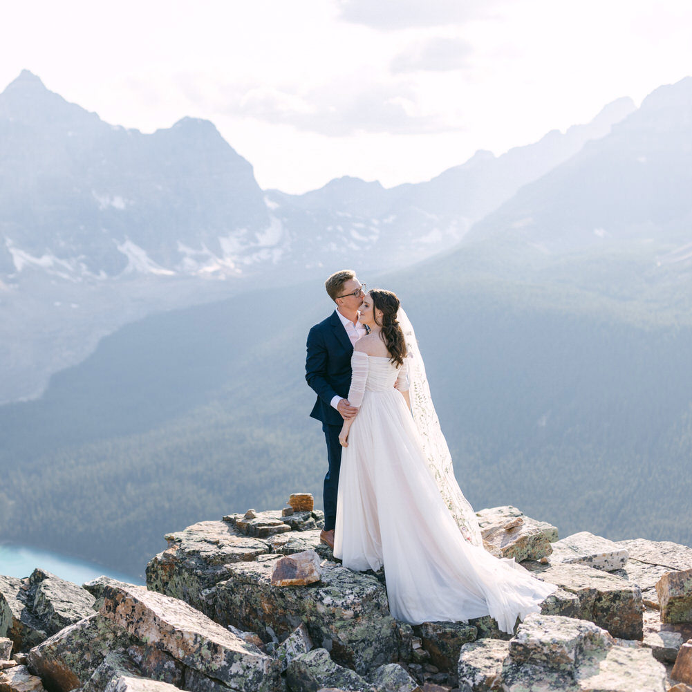 A bride and groom sharing a kiss on a rocky mountain summit with majestic peaks in the background.