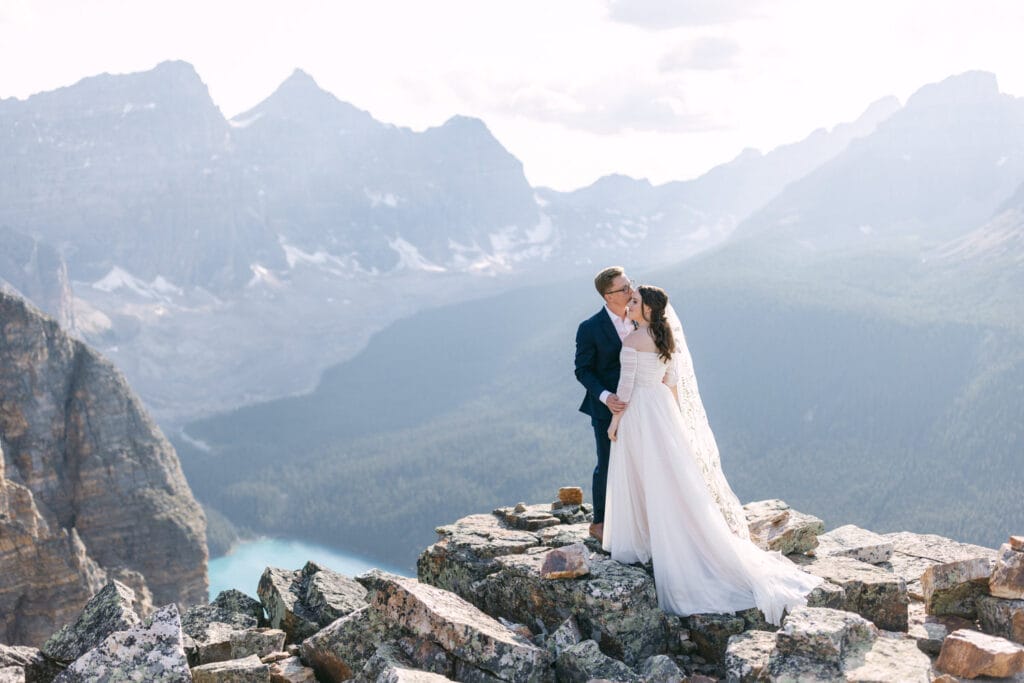 A wedding couple sharing a kiss on a rocky mountaintop with panoramic views of mountains and a turquoise lake.