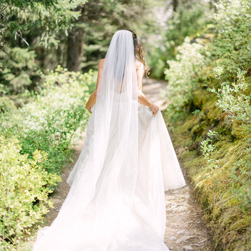 A bride in a white dress and veil walking down a forest trail.