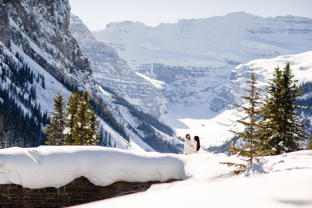 Two people hugging, partially obscured by a snow-covered ledge, with a breathtaking mountainous backdrop under a clear blue sky.