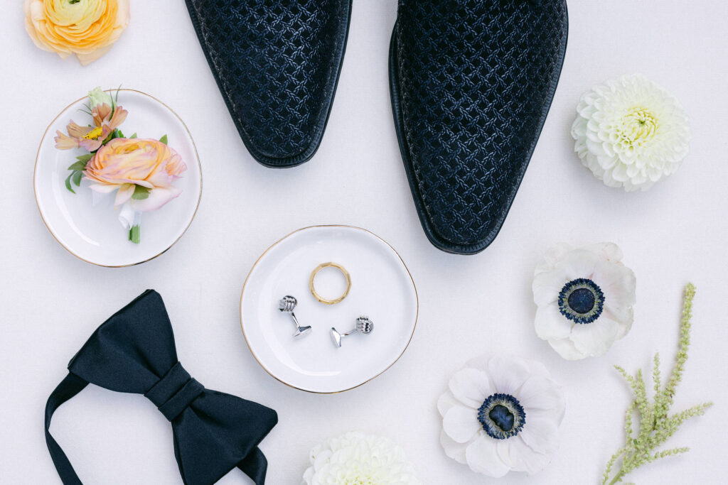 A flat lay of wedding accessories including black shoes, plates with a floral arrangement, a bow tie, and rings with cufflinks, complemented by fresh flowers on a light background.