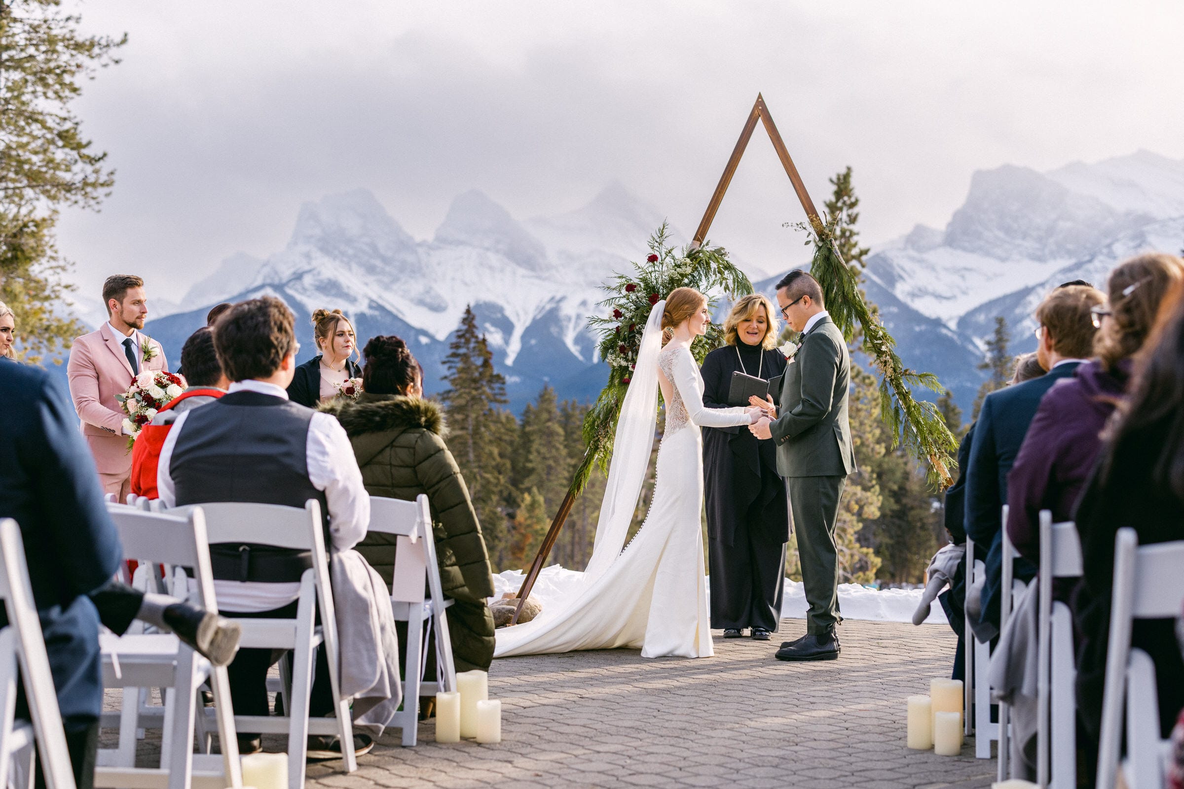 A bride and groom exchanging vows at an outdoor wedding ceremony with snowy mountains in the background and guests seated on either side.