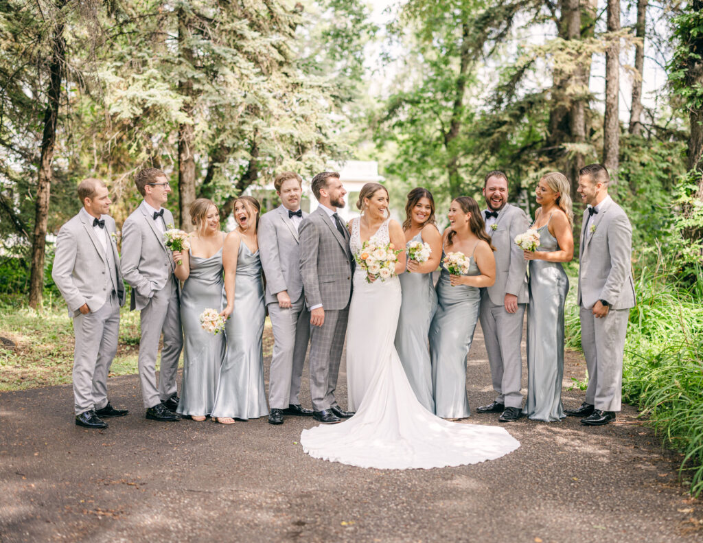 A bride and groom with their joyful wedding party, comprising bridesmaids and groomsmen, standing on a forest path, all dressed in formal wedding attire, sharing a laugh together.