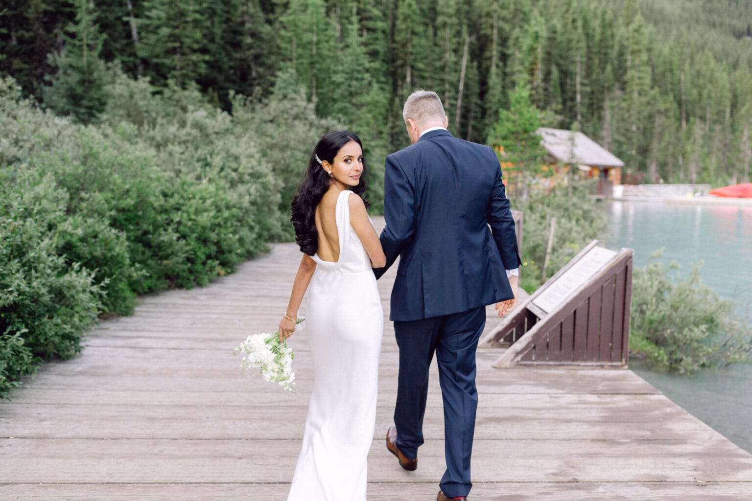 A bride and groom walking hand in hand along a wooden boardwalk near a lake, surrounded by lush greenery.