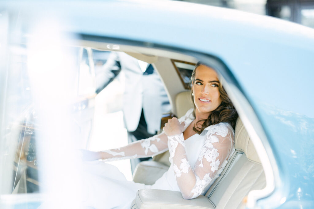 A bride in a lacy wedding dress with a beaming smile, seated inside a vintage car, captured through an overexposed shot.