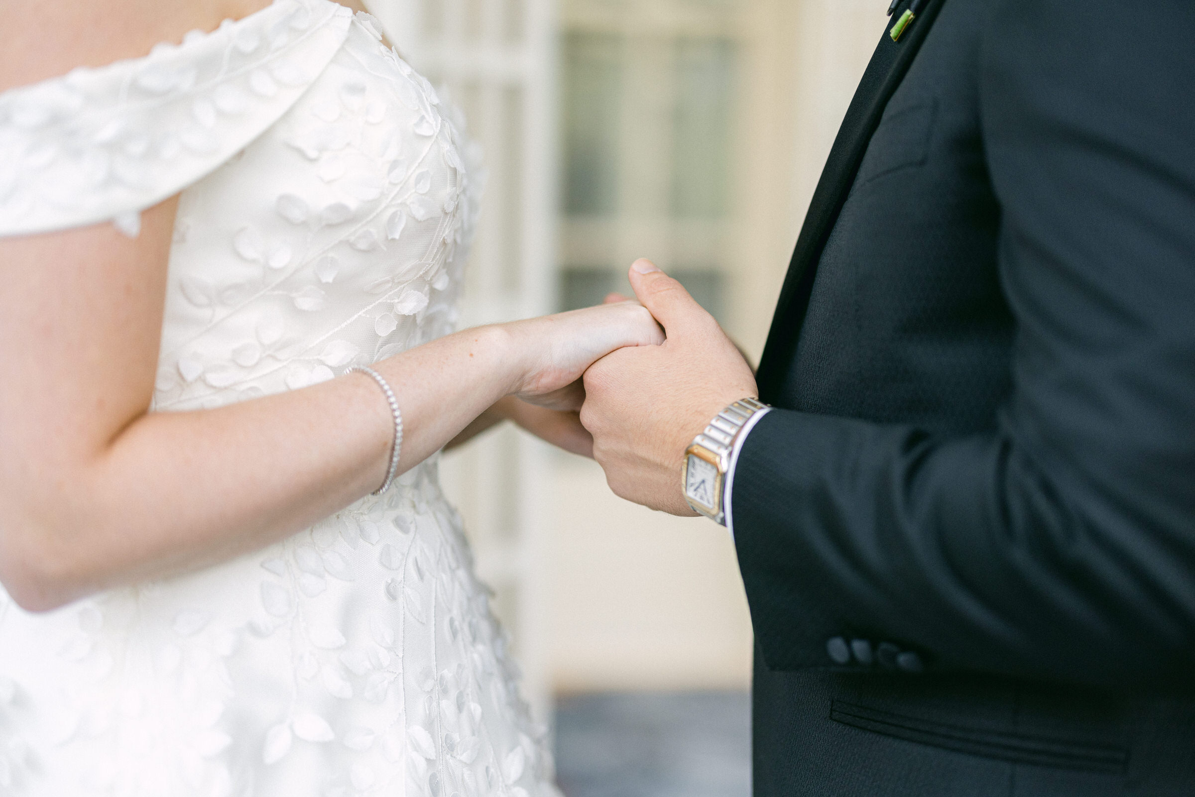 Close-up of a bride and groom holding hands on their wedding day, with focus on their hands and elegant attire.