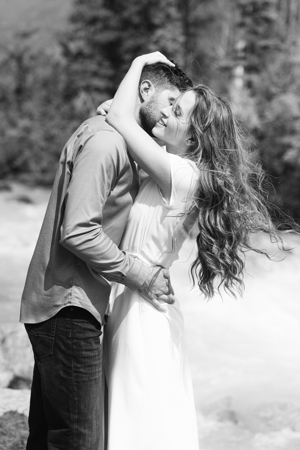 A black and white photograph of a couple embracing and sharing a loving moment outdoors with blurred natural scenery in the background.