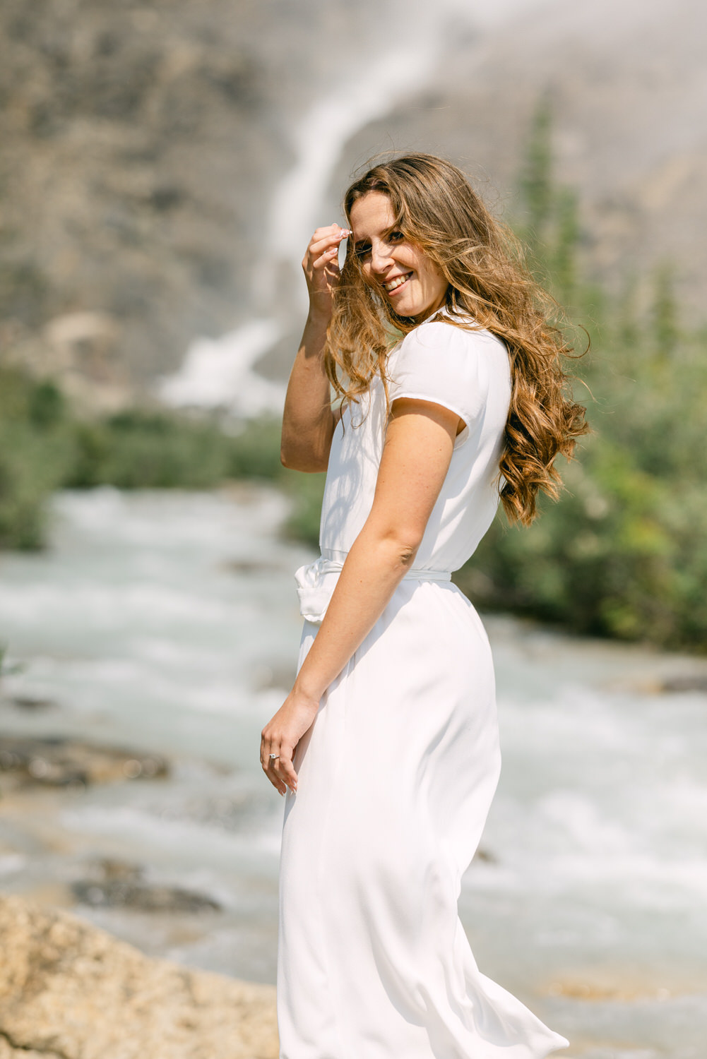 A smiling woman in a white dress standing by a river with a waterfall in the background.