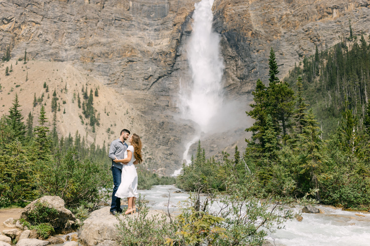 A couple hugging in front of a cascading waterfall surrounded by lush greenery.