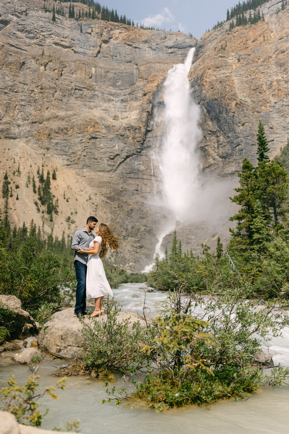 A couple hugging in front of a towering waterfall surrounded by rugged cliffs and greenery.