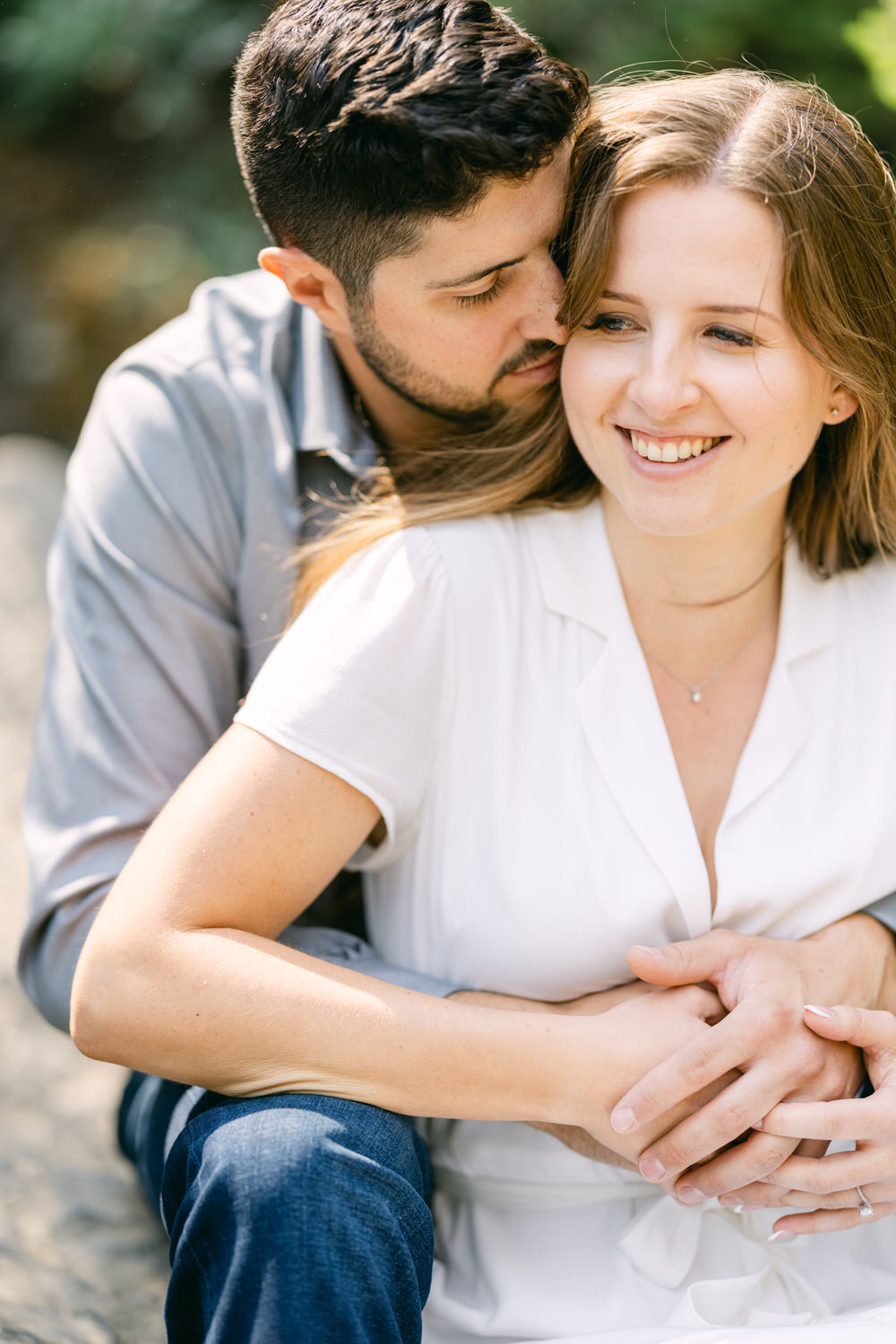 A man embracing a smiling woman from behind, both dressed casually outdoors.