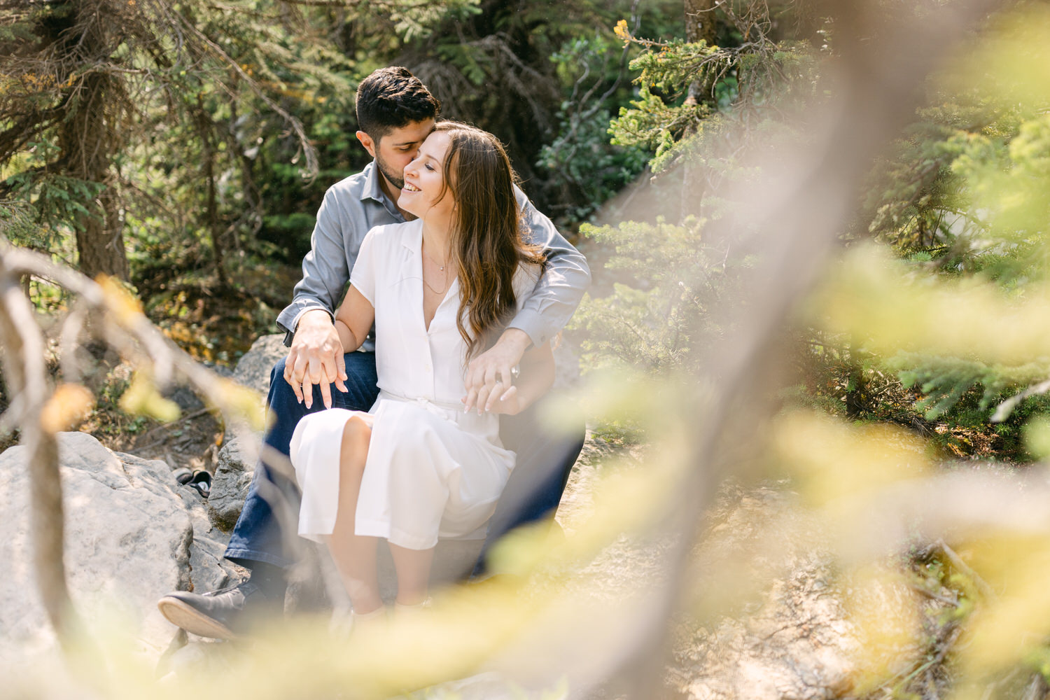 A couple embracing while sitting on rocks amidst lush greenery, with sunlight filtering through the foliage.