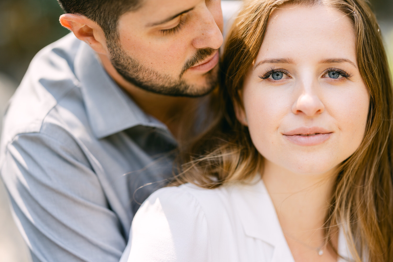 Close-up of a woman's face looking at the camera with a man leaning in close behind her, both dressed in light clothing, expressing affection.