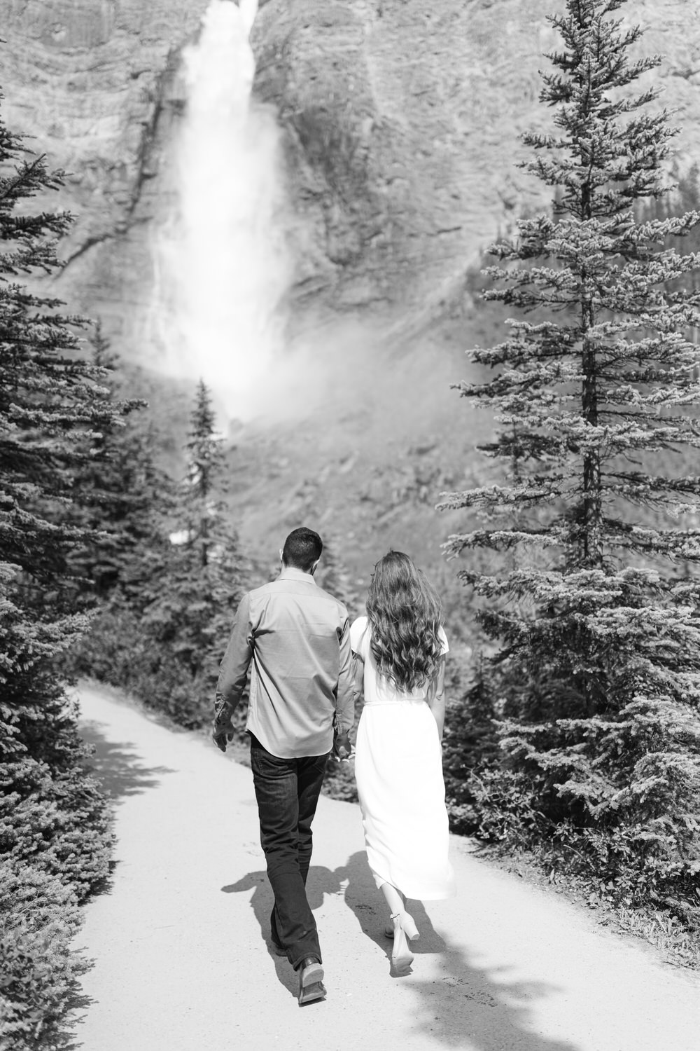 A black and white image of two people walking hand in hand towards a waterfall, surrounded by coniferous trees.