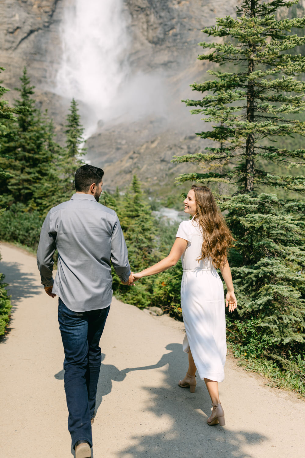 A man and woman holding hands while walking on a path, with a large waterfall in the background and surrounded by evergreen trees.