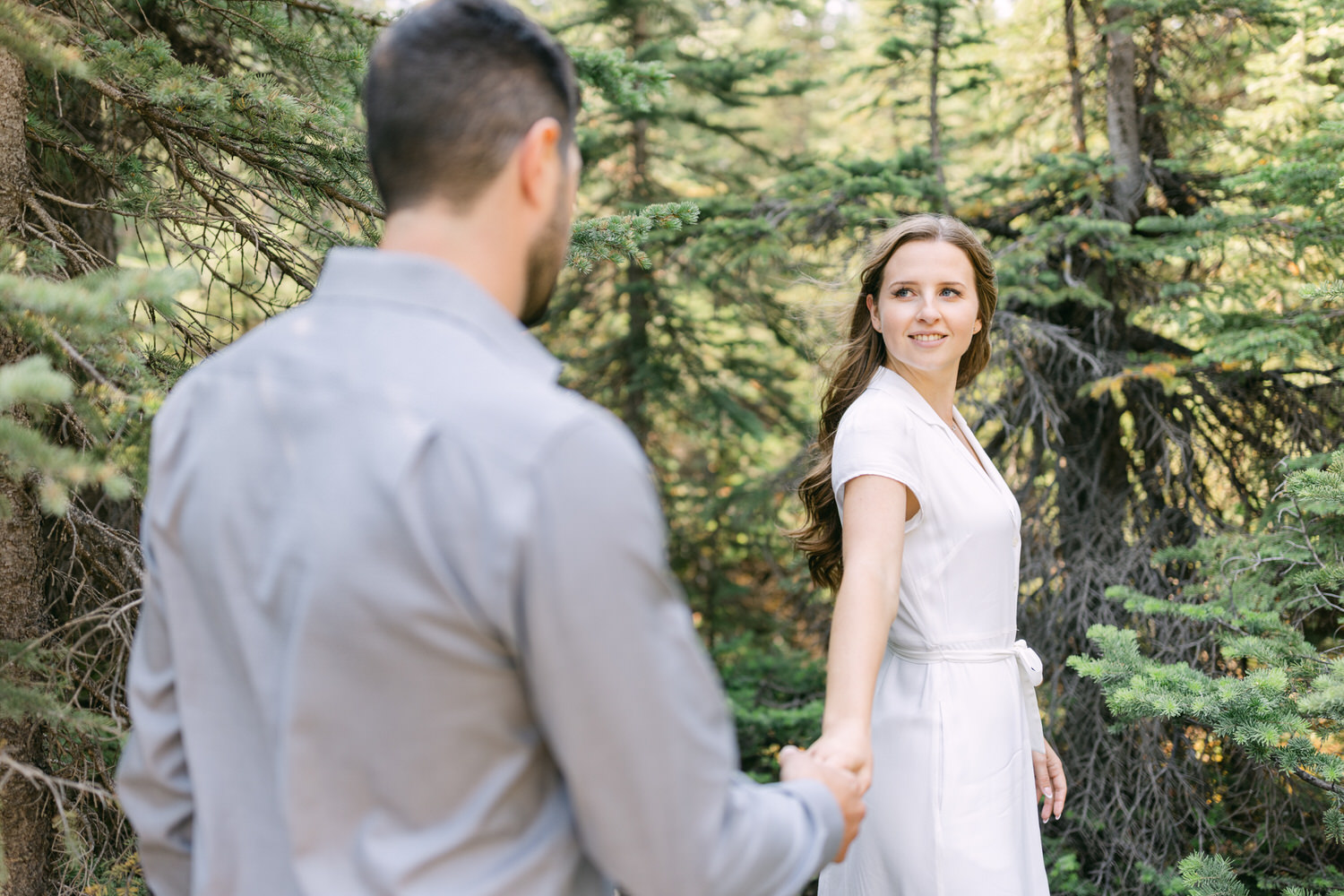 A woman in a white dress holding hands with a man in a gray shirt, looking back and smiling amidst a lush green forest setting.