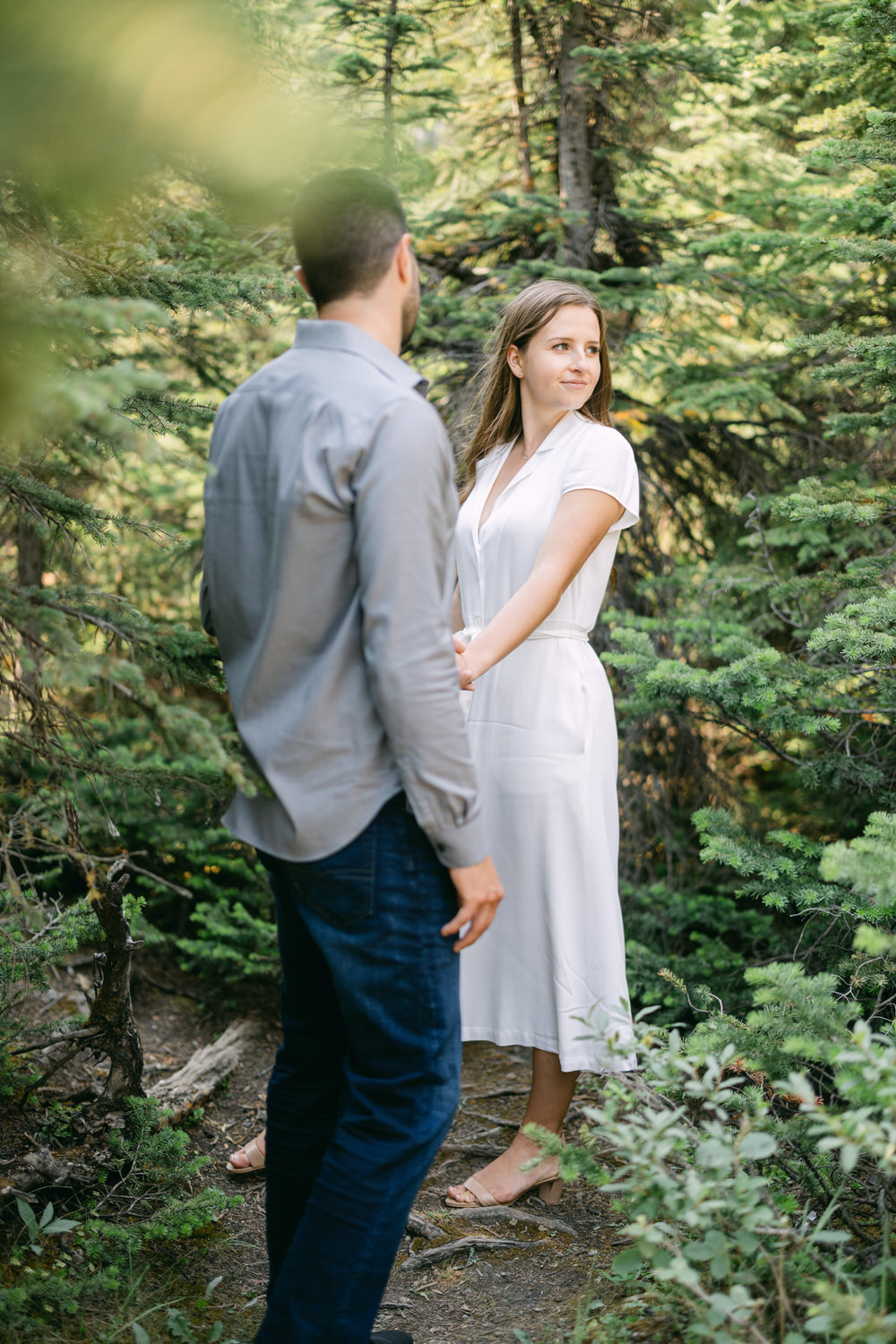 A man and woman standing in a forest, with the woman looking over her shoulder at the camera while holding hands with the man whose back is facing the camera.