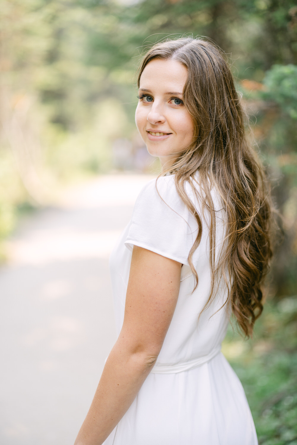 A woman in a white dress looking over her shoulder with a soft smile against a blurred green natural background.