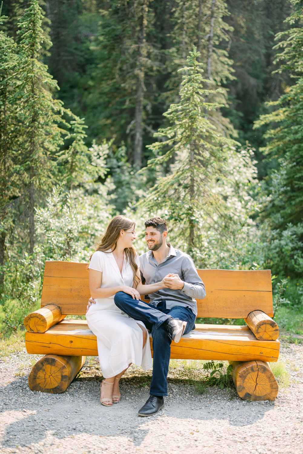 A couple sitting closely on a wooden bench in a forest, smiling and looking at each other with green trees in the background.