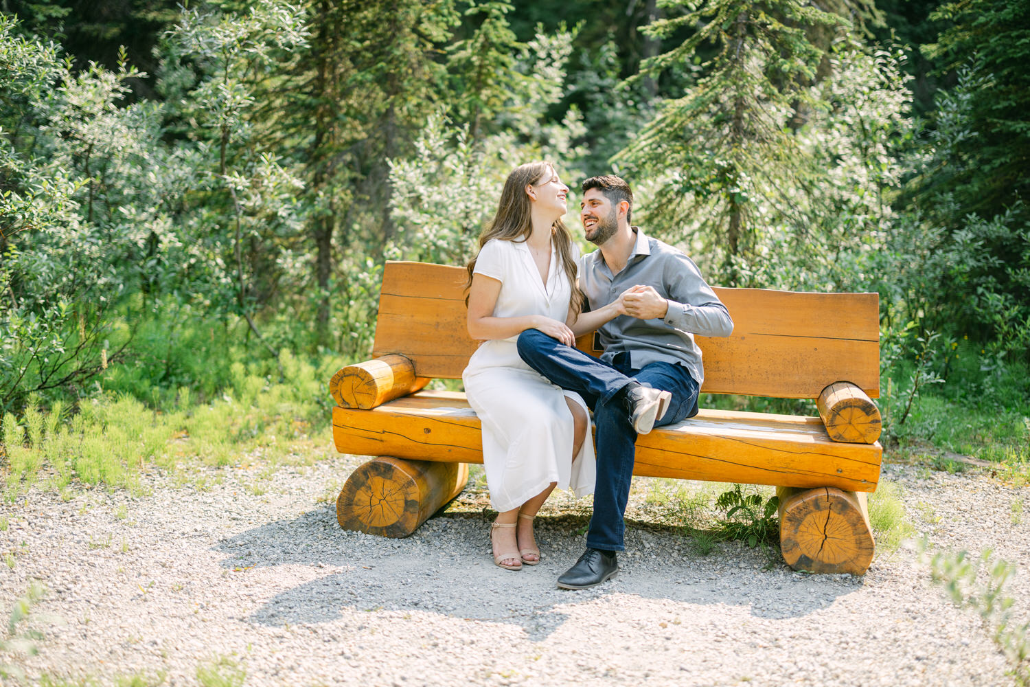 A man and woman sitting closely and smiling at each other on a wooden bench in a lush green park setting.