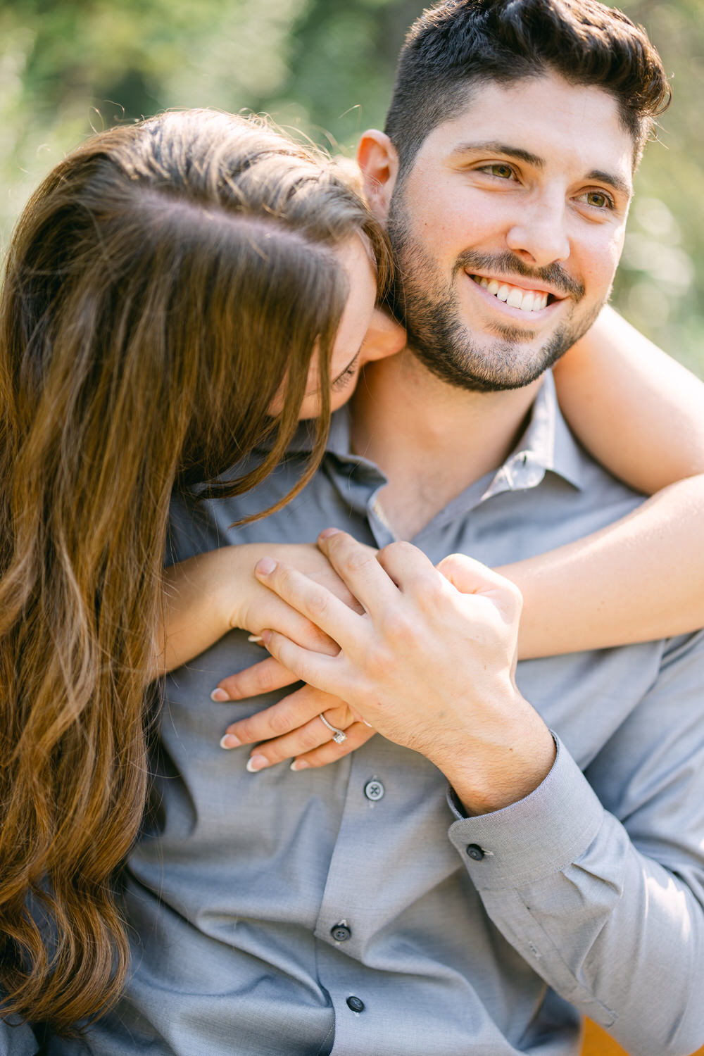 A man smiling at the camera while a woman rests her head on his shoulder, both bathed in natural light.