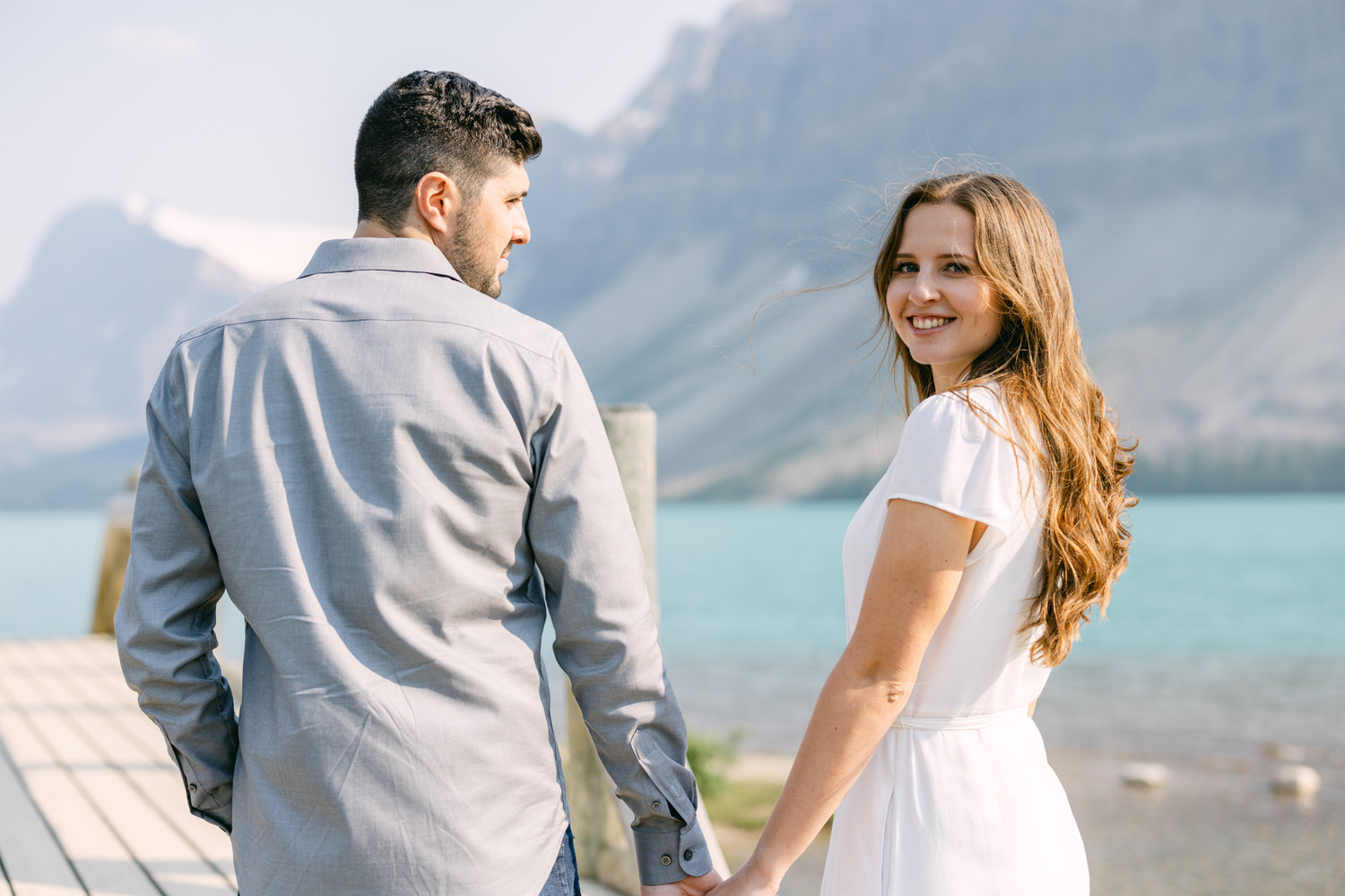 A couple holding hands by a turquoise lake with mountains in the background, man facing away and woman facing the camera with a smile