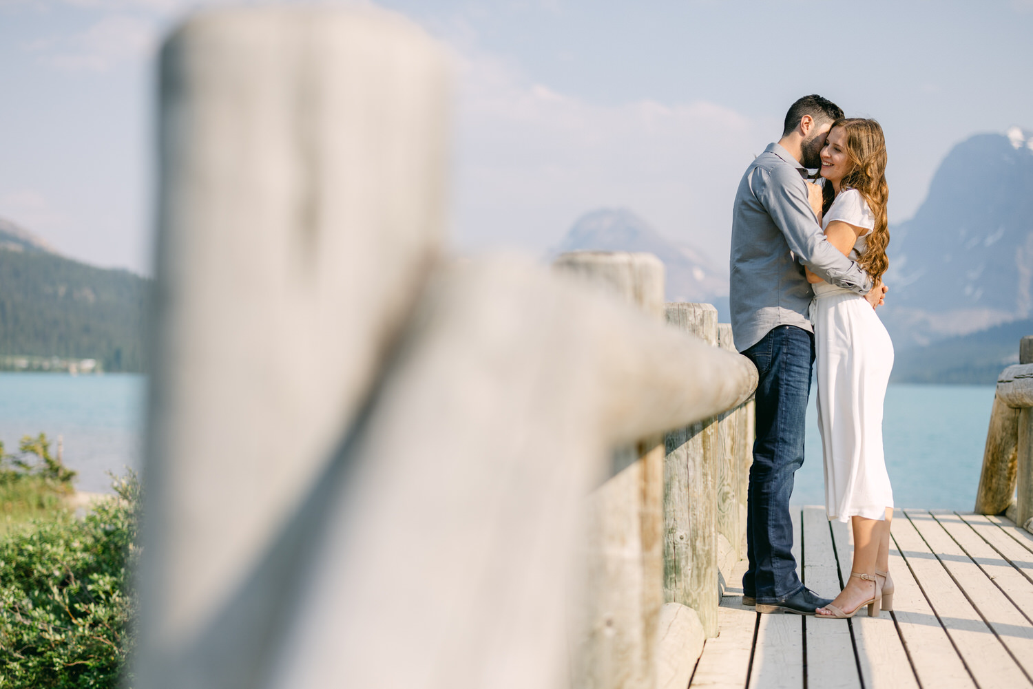 A couple hugging each other on a wooden dock by a lake with mountains in the background