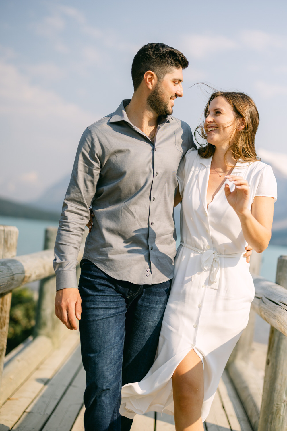 A smiling couple walking hand in hand on a wooden dock with a scenic lake and sky in the background.