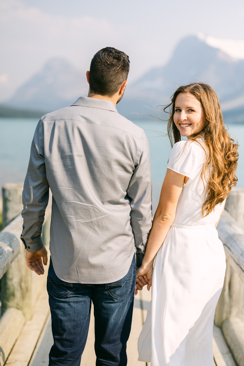 Two people holding hands while walking on a wooden pier, with a woman looking back and smiling, against a backdrop of mountains and a clear sky.