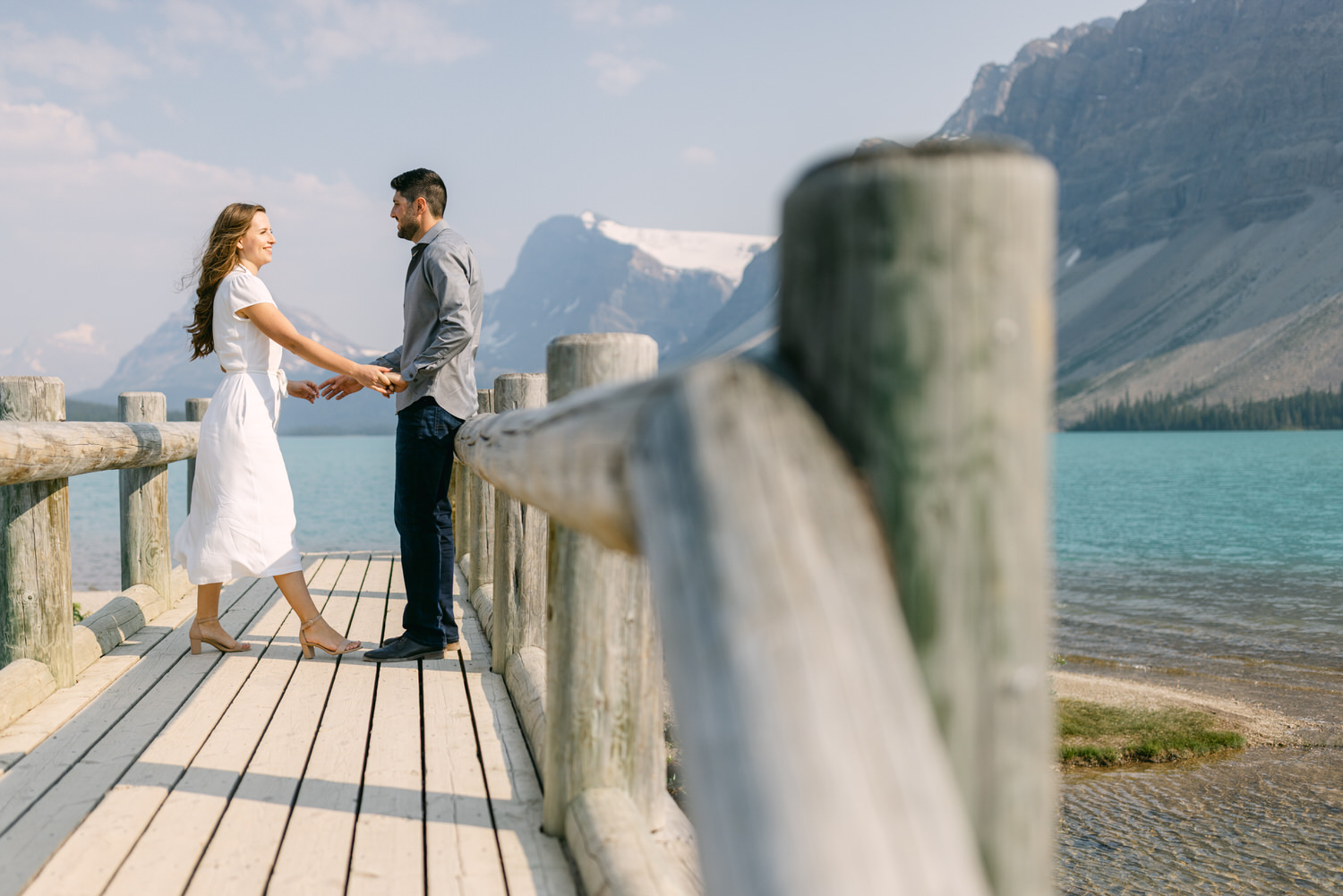 A couple holding hands on a wooden pier overlooking a turquoise lake with mountains in the background