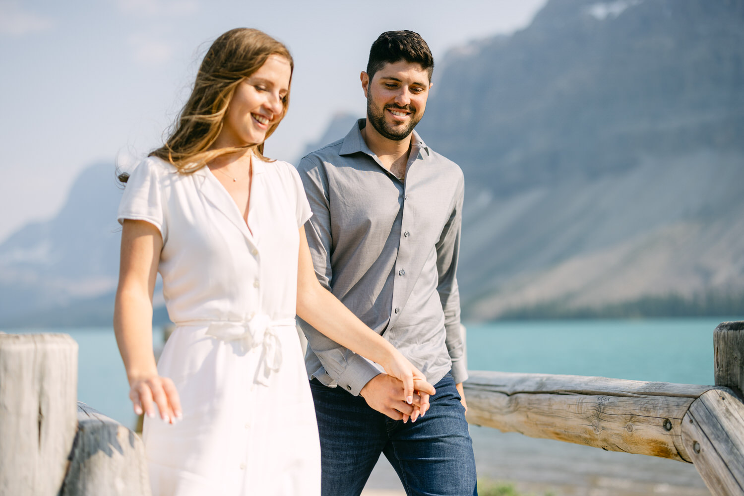 A man and woman holding hands and smiling while walking together against a backdrop of a clear blue mountain lake and hills.