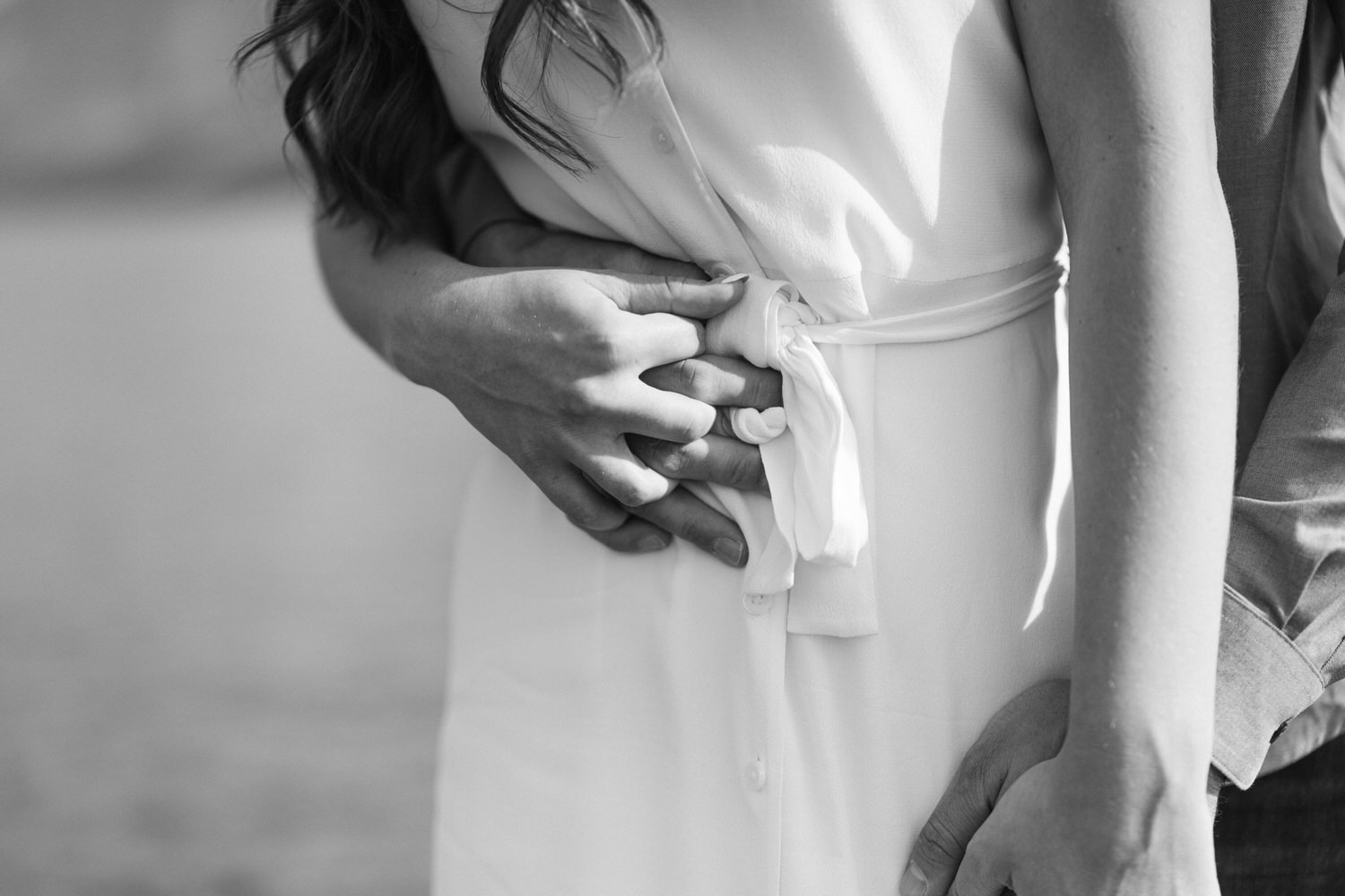 Black and white image of a couple in a close embrace, focusing on their hands tenderly clasped around the woman's waist.