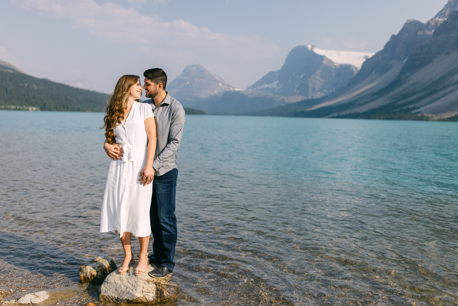 A man and woman embracing on a rock at the edge of a serene mountain lake with mountains in the background.