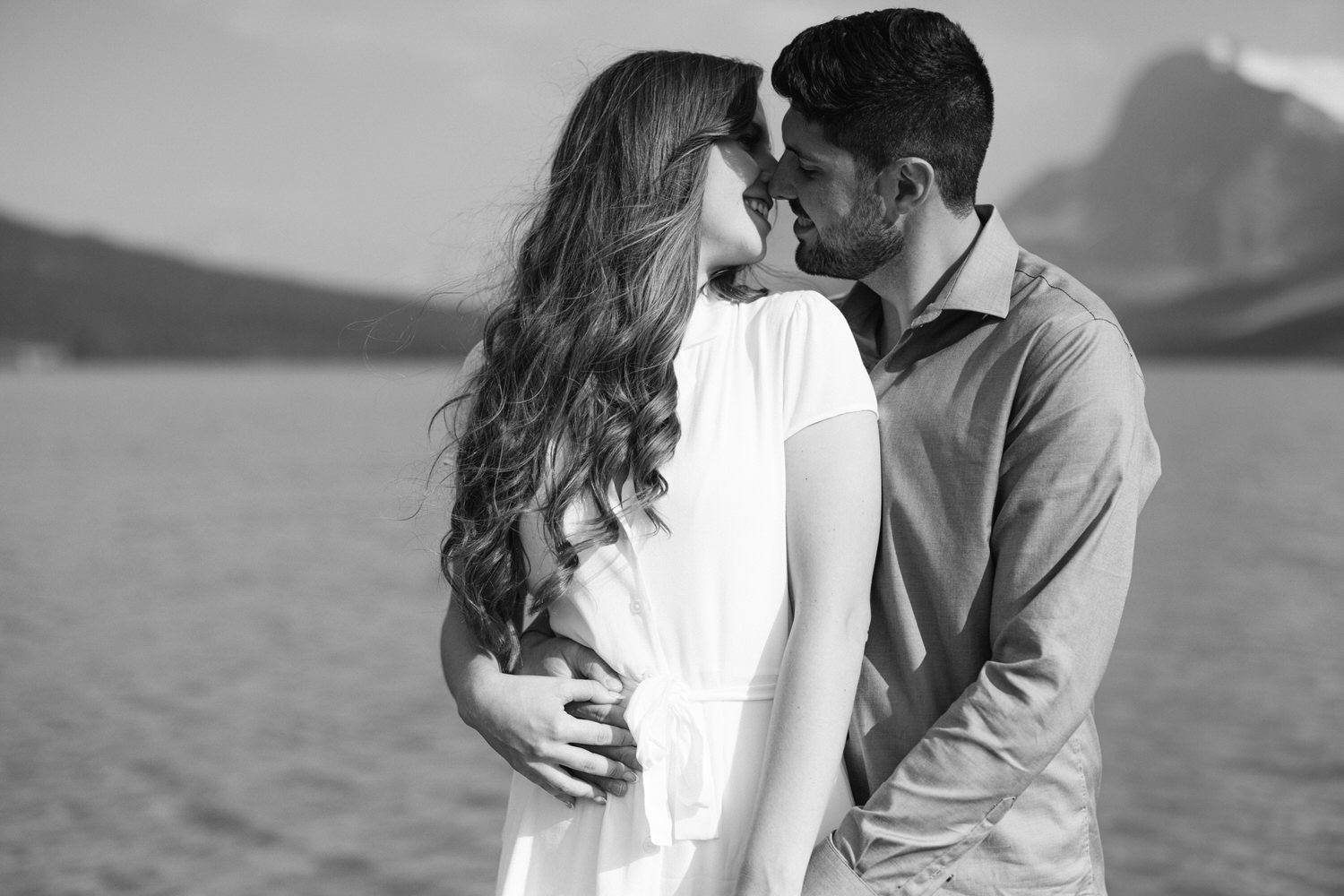 Black and white image of a couple embracing and touching foreheads affectionately near a body of water, with mountains in the soft-focus background.