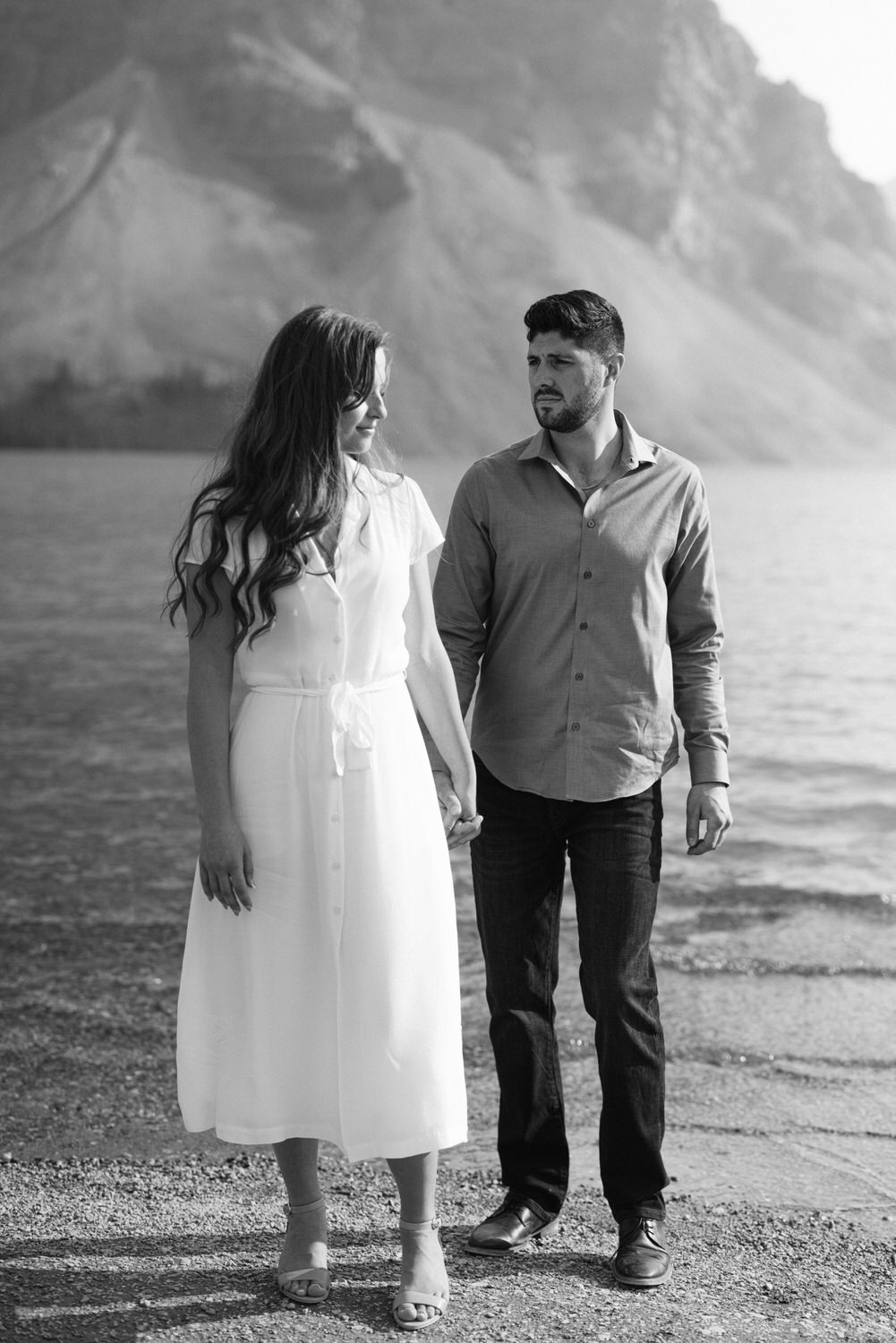 A man and woman holding hands and looking at each other by a lake with mountains in the background, in black and white.